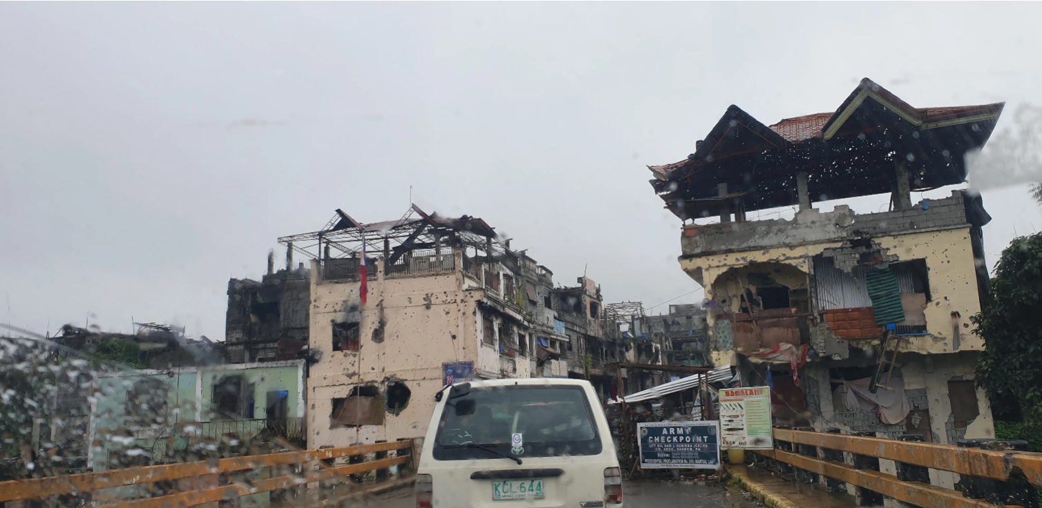 a military checkpoint on a bridge in a war-damaged Marawi City, with heavily damaged buildings showing bullet holes and structural destruction under overcast weather conditions.