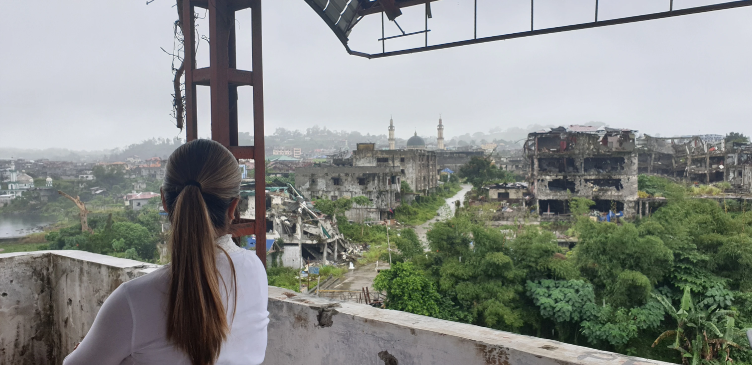 Woman observing a devastated urban landscape with destroyed buildings, greenery, and mosque structures in the distance from an elevated viewpoint, reflecting a post-conflict scene in Marawi.
